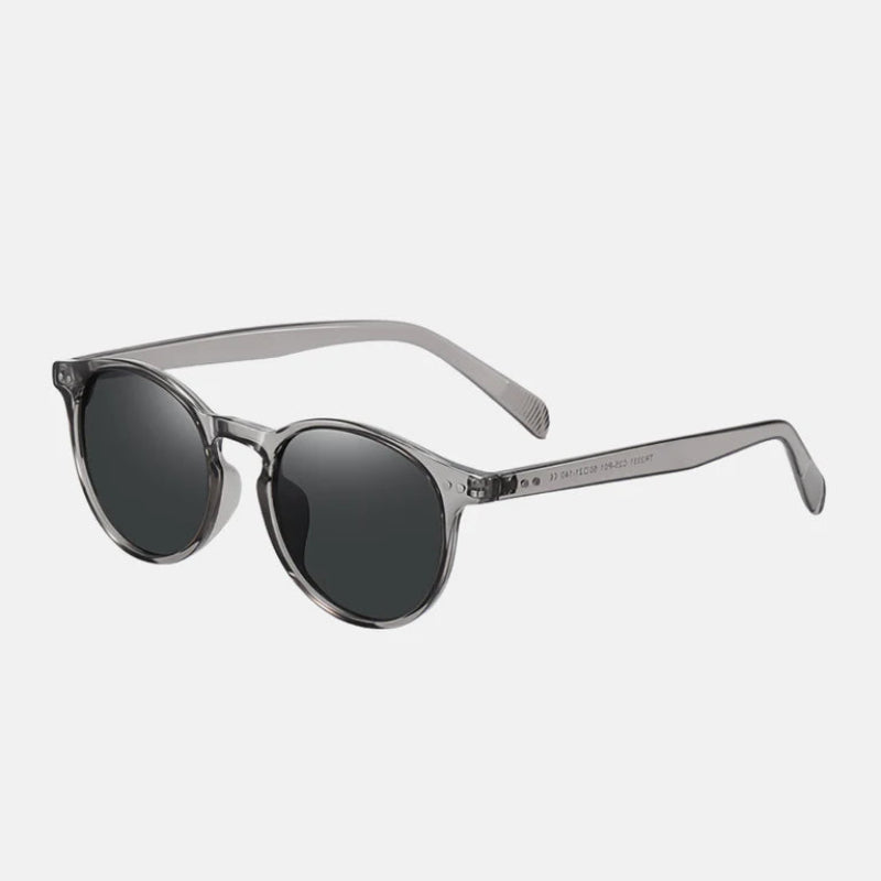 Sunglasses Lico Grey – stylish round-frame sunglasses in a transparent grey acetate design, with dark grey polarized lenses. The temples are straight and slim, maintaining the same transparent grey tone, with visible hinge detailing and engraved model specifications on the inner arm. Shot at a 45-degree angle to highlight the frame depth and lens color, on a white background. www.zenluk.com